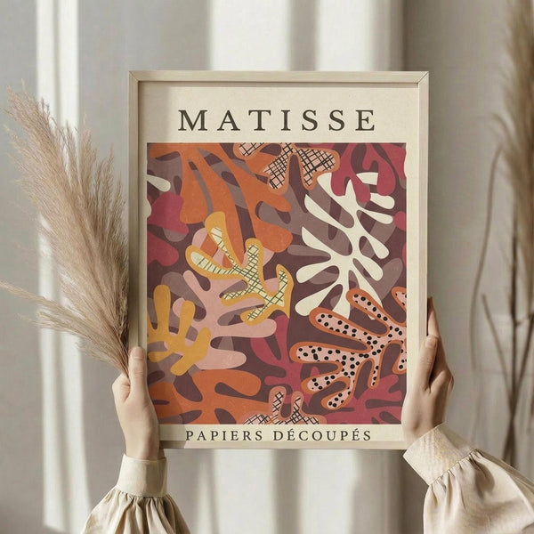 May include: Person holding framed Matisse-inspired art print featuring abstract cut-out paper shapes in warm autumn colors - orange, burgundy, yellow, and cream - with crosshatch patterns and organic flowing forms. Light wood frame against neutral curtained background, dried pampas grass visible on left side.