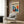 May include: Framed Matisse-inspired modernist poster hanging on wood-paneled wall above beige upholstered armchair with geometric pillow. Artwork features silhouetted dancing figure surrounded by vibrant blue, orange, and yellow abstract shapes. Room setting includes brass floor lamp and side table with open book.