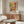 May include: Colorful Matisse-style poster in wooden frame hanging above mid-century walnut credenza in modern living room. Artwork features vibrant interior scene with plants, patterned chair, and geometric shapes in yellow, coral, and blue tones. Room styled with cream curved chair, glass coffee table, and brass floor lamp.