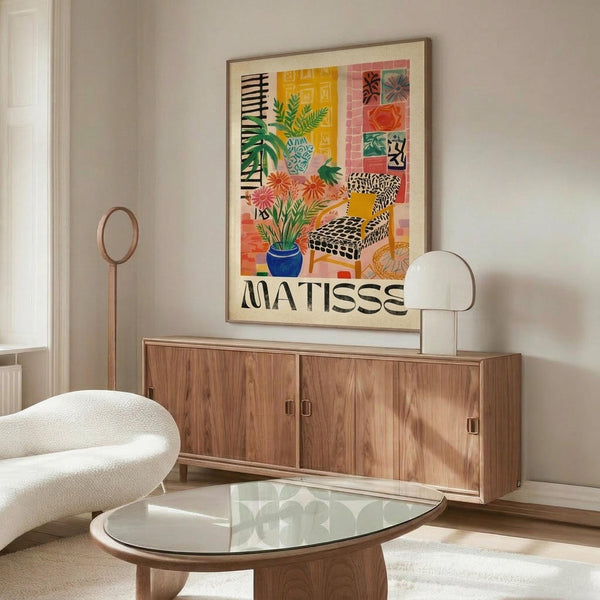 May include: Colorful Matisse-style poster in wooden frame hanging above mid-century walnut credenza in modern living room. Artwork features vibrant interior scene with plants, patterned chair, and geometric shapes in yellow, coral, and blue tones. Room styled with cream curved chair, glass coffee table, and brass floor lamp.