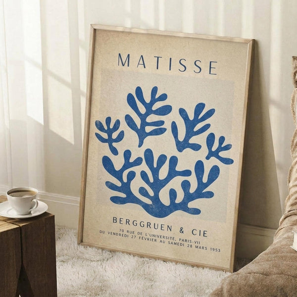 May include: Framed art print leaning against white wall corner on textured white rug. Poster features blue coral-like branching design on cream background with "MATISSE" text at top and gallery information below. Light wood frame, displayed next to wooden side table with white coffee cup and saucer. Soft natural lighting creates gentle shadows.