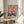 May include: Colorful floral art print in wooden frame displayed on round wooden side table next to beige upholstered chair. Vibrant bouquet illustration features pink, orange, red and yellow blooms with blue-green foliage in painterly style. Artist name appears at bottom in black text. Bright natural lighting creates soft shadows on neutral wall background with palm plant visible at left edge.