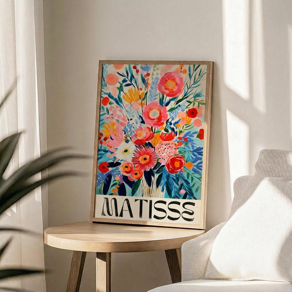May include: Colorful floral art print in wooden frame displayed on round wooden side table next to beige upholstered chair. Vibrant bouquet illustration features pink, orange, red and yellow blooms with blue-green foliage in painterly style. Artist name appears at bottom in black text. Bright natural lighting creates soft shadows on neutral wall background with palm plant visible at left edge.