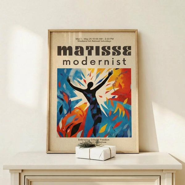 May include: Framed poster displayed on light wood console table against white wall. Poster features bold modernist design with silhouetted figure in dance pose, arms raised, surrounded by vibrant blue, orange, and yellow abstract shapes. Text reads "matisse modernist" with event details above. Small white gift box with twine sits nearby on table surface.