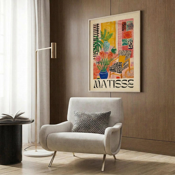 May include: Colorful Matisse-style poster in light wood frame hanging on wood-paneled wall above beige upholstered chair with geometric pillow. Artwork features vibrant collage of tropical plants, patterned chair, and decorative elements in yellows, oranges, and greens. Modern room setting with brass floor lamp and side table.