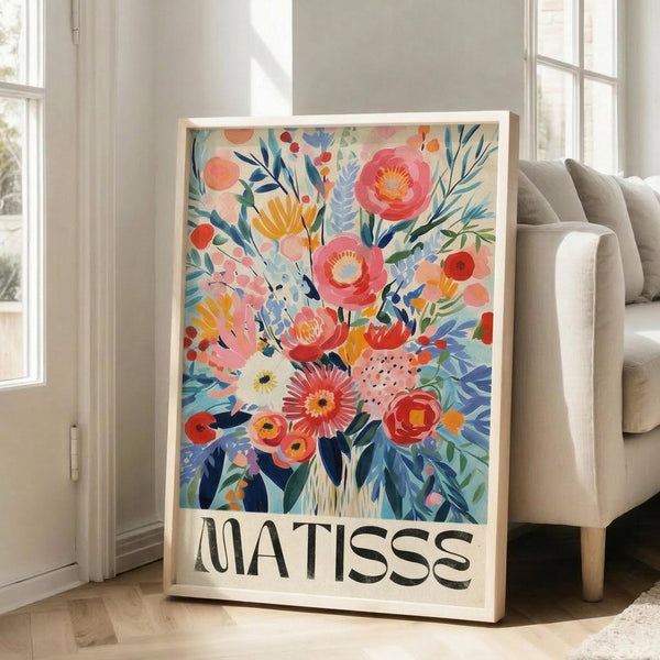 May include: Large framed art print featuring vibrant watercolor-style floral bouquet with pink, coral, orange and blue flowers against cream background, displayed on light wooden floors next to beige upholstered chair in bright room with white walls and windows. The botanical artwork shows loose, painterly brushstrokes in a modern illustrative style with "MATISSE" text at bottom in bold black letters.