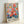 Colorful floral artwork in light wood frame leaning against white wall. Vibrant bouquet features pink peonies, coral flowers, yellow blooms, and blue-green foliage in loose, painterly style. Artist name "MATISSE" appears in black text at bottom of cream-colored background. Frame casts soft shadow on wall surface.