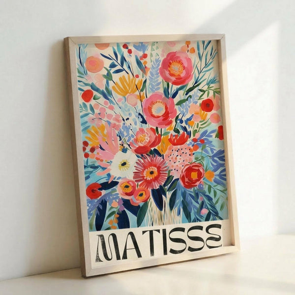 Colorful floral artwork in light wood frame leaning against white wall. Vibrant bouquet features pink peonies, coral flowers, yellow blooms, and blue-green foliage in loose, painterly style. Artist name "MATISSE" appears in black text at bottom of cream-colored background. Frame casts soft shadow on wall surface.