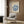 May include: Beige armchair with geometric patterned pillow, gold floor lamp, dark round side table with open book, and framed Matisse exhibition poster featuring blue coral-like botanical forms on cream background. Modern living room setting with wood panel wall, white curtains, and light wooden flooring.