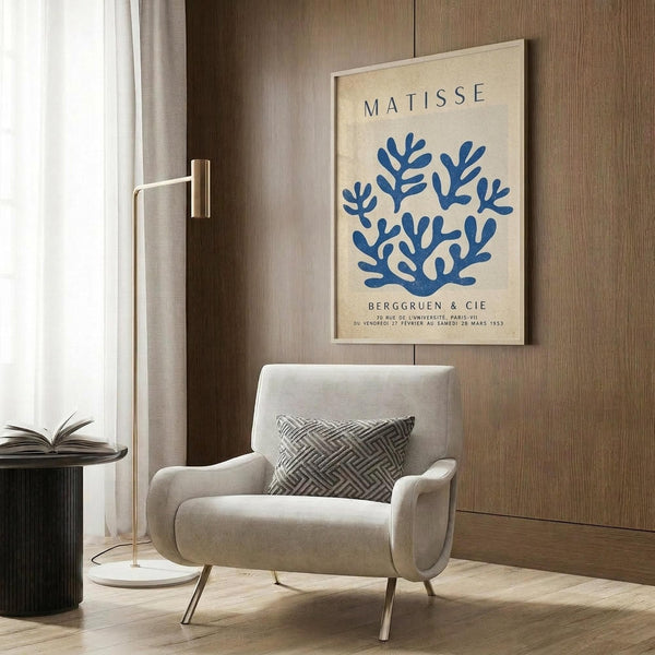 May include: Beige armchair with geometric patterned pillow, gold floor lamp, dark round side table with open book, and framed Matisse exhibition poster featuring blue coral-like botanical forms on cream background. Modern living room setting with wood panel wall, white curtains, and light wooden flooring.
