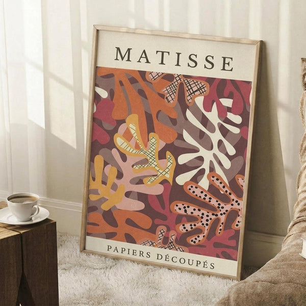 May include: Framed Matisse-style poster with abstract coral and botanical cutout shapes in warm oranges, reds, and browns, leaning against white wall corner next to small wooden stool with white coffee cup, beige textured rug below, natural lighting creating soft shadows