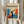 May include: Hands holding a framed poster featuring a black silhouette figure with raised arms against vibrant blue and orange abstract brushstrokes in Matisse-inspired style. The cream-colored poster has dark text at top and bottom. Person wears beige clothing and holds dried pampas grass, with soft natural lighting and neutral curtained background.