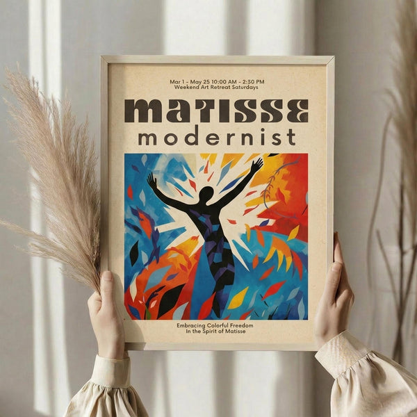 May include: Hands holding a framed poster featuring a black silhouette figure with raised arms against vibrant blue and orange abstract brushstrokes in Matisse-inspired style. The cream-colored poster has dark text at top and bottom. Person wears beige clothing and holds dried pampas grass, with soft natural lighting and neutral curtained background.