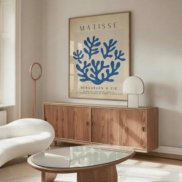 May include: Matisse exhibition poster with blue coral-like design on cream background, displayed in modern living room with wooden sideboard, white curved chair, glass coffee table, and contemporary lamp. Poster shows "BERGGRUEN & CIE" gallery text and Paris exhibition details from 1953.