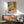 May include: Colorful Matisse-style poster print featuring tropical plants, patterned chair, and vibrant geometric shapes in yellow, pink, green, and orange tones, displayed in wooden frame above modern credenza with white ceramic lamp and sage green mid-century armchair in sunlit room with white walls.