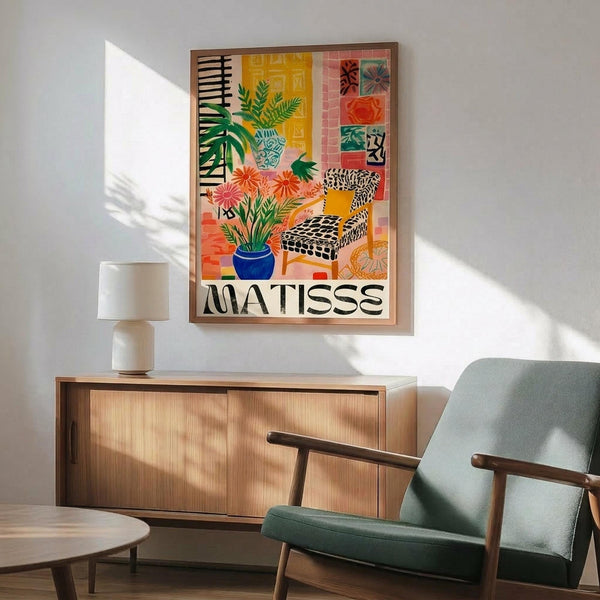 May include: Colorful Matisse-style poster print featuring tropical plants, patterned chair, and vibrant geometric shapes in yellow, pink, green, and orange tones, displayed in wooden frame above modern credenza with white ceramic lamp and sage green mid-century armchair in sunlit room with white walls.