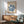 May include: Framed Matisse exhibition poster featuring blue coral-like cutout design on cream background, displayed above wooden credenza with table lamp, next to sage green mid-century modern armchair with wooden arms in sunlit room with white walls and geometric shadows.
