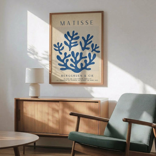 May include: Framed Matisse exhibition poster featuring blue coral-like cutout design on cream background, displayed above wooden credenza with table lamp, next to sage green mid-century modern armchair with wooden arms in sunlit room with white walls and geometric shadows.