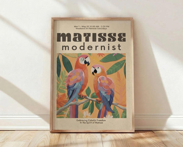 May include: Framed poster with colorful parrots perched on branches surrounded by tropical leaves in Matisse-inspired cut-out style. Features vibrant oranges, pinks, blues, and greens against warm yellow background. Text reads "matisse modernist" and "Embracing Colorful Freedom In the Spirit of Matisse" with event details. Natural wood frame displayed on light wooden floor against white wall with soft shadows.