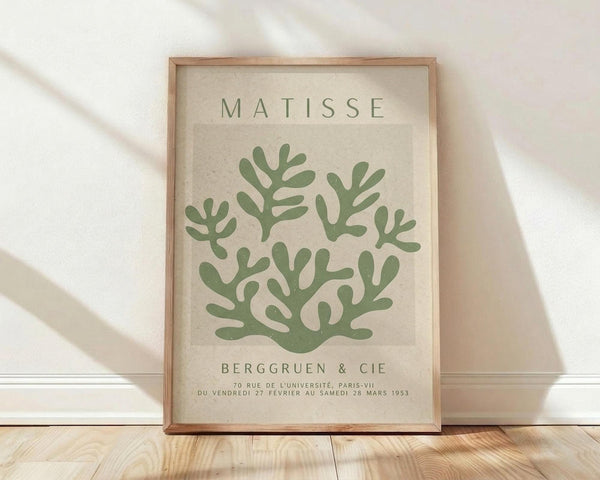 May include: Framed art print with beige background featuring green coral-like botanical shapes in Matisse's cut-out style. Text reads "MATISSE" at top and "BERGGRUEN & CIE" with French exhibition details below. Natural wood frame sits on light wooden floor against white wall with soft natural lighting creating gentle shadows.