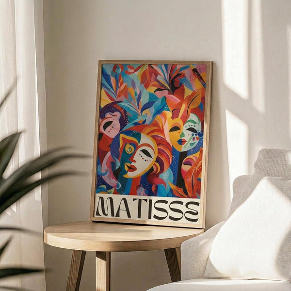 May include: Colorful abstract art poster in thin gold frame displayed on round wooden side table next to beige upholstered chair. Artwork features vibrant red, blue, yellow and orange stylized faces and foliage with "MATISSE" text at bottom. Soft natural lighting creates shadows on white wall, with green plant leaves visible in corner.