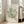 May include: Framed botanical poster with green coral-like shapes on cream background, displayed in bright living room corner next to beige sofa with pillows, natural sunlight streaming through white-framed windows onto light wooden floors.