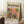 May include: Framed poster with colorful parrots perched on branches surrounded by tropical leaves, displayed against white paneled wall on textured white rug. Natural wood frame contains vintage-style art print with orange and yellow background. Small wooden side table with white coffee cup visible on left. Cozy home setting with beige throw blanket draped nearby.