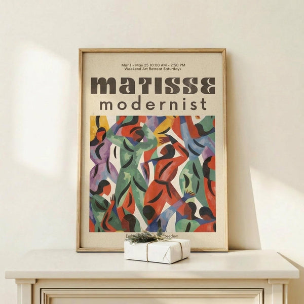 May include: Framed exhibition poster displaying abstract modernist artwork with flowing organic shapes in vibrant colors including green, orange, purple, and blue against cream background. The artwork features curved, interlocking forms resembling dancing figures. Poster sits on light wood surface next to small white gift box with twine and greenery. Natural lighting creates soft shadows on white wall behind.