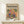 May include: Framed poster displaying two colorful parrots perched on a branch surrounded by tropical leaves in Matisse-inspired modernist style, featuring vibrant oranges, blues, and greens against a warm yellow background. Light wood frame sits on pale wooden surface beside small white wrapped gift with twine and greenery sprig against cream wall.