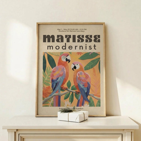 May include: Framed poster displaying two colorful parrots perched on a branch surrounded by tropical leaves in Matisse-inspired modernist style, featuring vibrant oranges, blues, and greens against a warm yellow background. Light wood frame sits on pale wooden surface beside small white wrapped gift with twine and greenery sprig against cream wall.