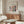 May include: Matisse-inspired art poster in thin wooden frame mounted above walnut credenza in minimalist living room. Poster features colorful window scene with green vase, pink and red flowers, blue accents on cream background. Modern white curved chair, glass oval coffee table, and white table lamp complete the Scandinavian-style interior with white walls and natural lighting.