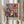 May include: Two hands holding a framed colorful art print in a light wooden frame against white curtains, with dried pampas grass visible to the left. The artwork features vibrant abstract faces and masks in blues, reds, oranges, and yellows with flowing organic shapes and foliage elements in a modern artistic style reminiscent of paper cutouts.