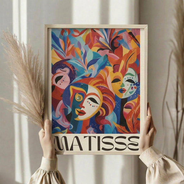 May include: Two hands holding a framed colorful art print in a light wooden frame against white curtains, with dried pampas grass visible to the left. The artwork features vibrant abstract faces and masks in blues, reds, oranges, and yellows with flowing organic shapes and foliage elements in a modern artistic style reminiscent of paper cutouts.