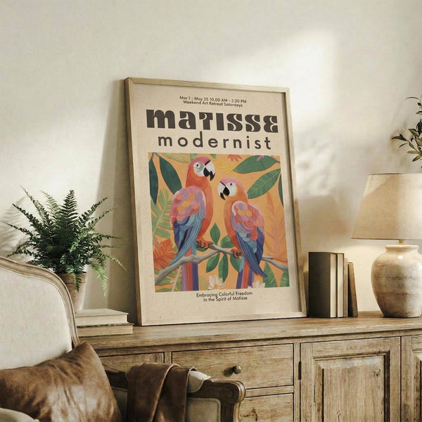May include: Framed poster featuring two colorful parrots perched on a branch surrounded by tropical leaves in Matisse-inspired style with vibrant pinks, blues, and greens against yellow background. Shows exhibition details at top and "Embracing Colorful Freedom In the Spirit of Matisse" text below. Display shows poster leaning on rustic wooden sideboard with books, ceramic vase, and fern plant, with cream upholstered chair visible in cozy living room setting.