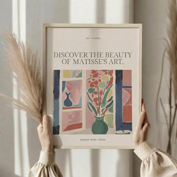 May include: Hands holding a framed art poster featuring a Matisse-inspired interior scene with a green vase of red flowers on a windowsill, geometric shapes in pink, coral, and blue tones, and a small dark vase visible through the window. Light wooden frame against neutral curtained background with dried pampas grass visible on the left side.