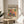 May include: A framed poster featuring two colorful parrots perched on branches surrounded by tropical foliage in vibrant oranges, greens, and blues. The artwork displays "matisse modernist" text at the top with "Embracing Colorful Freedom In the Spirit of Matisse" below. The print sits in a thin wooden frame on a round wooden side table next to a beige upholstered chair, with natural sunlight casting shadows across the neutral-toned room and a plant visible in the corner.