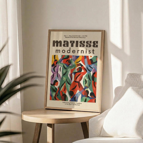 May include: Framed exhibition poster with "Matisse Modernist" text and vibrant abstract artwork featuring flowing organic shapes in red, green, blue, purple, and orange. Displayed on light wood round side table next to beige upholstered chair with plant leaves visible. Natural sunlight creates shadows on white wall background.