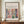 May include: Framed art poster with Matisse-inspired illustration of flowers in a green vase by a window, featuring coral, pink, green and blue colors in cut-out style. Poster displays against cream wall on wooden sideboard next to white ceramic vase and stacked books in minimalist home setting.