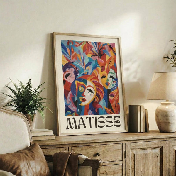 May include: Colorful abstract art print featuring stylized faces with decorative masks surrounded by vibrant foliage in blues, reds, oranges and yellows. The framed poster displays "MATISSE" text below the artwork and sits on a rustic wooden sideboard alongside books, a ceramic vase, and fern plant in a neutral-toned living room setting.