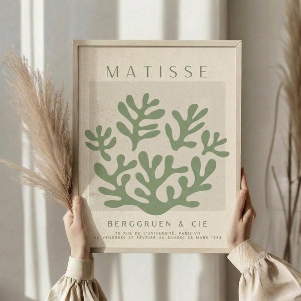 May include: Person in cream ruffled sleeves holding framed botanical art print featuring green coral-like shapes on beige background, with dried pampas grass in left hand, against soft white curtained window backdrop in minimalist neutral setting