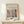 May include: Matisse-inspired art poster in natural wood frame displaying colorful interior scene with green vase of red flowers by window with blue frames, displayed on light wood surface next to small wrapped gift with twine and greenery sprig against cream wall