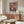 May include: Colorful abstract art poster featuring stylized faces and foliage in vibrant blues, reds, oranges, and yellows, displayed in thin gold frame above wooden mid-century modern credenza. Room shows white curved chair, glass oval coffee table, marble table lamp, and neutral walls with natural lighting.