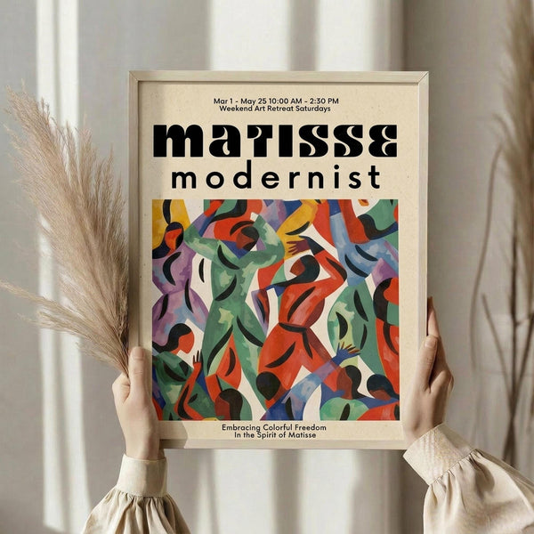 May include: Person holding a framed modernist exhibition poster featuring abstract colorful cut-out shapes in red, green, blue, and purple. The cream-colored poster displays bold black typography and vibrant geometric forms reminiscent of paper collage art. Dried pampas grass visible beside the light wood frame against soft neutral curtains and natural lighting.