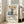 May include: Framed abstract art print featuring colorful still life with blue ceramic vases, orange and red fruits, yellow lemon, and botanical elements in Matisse-inspired style. Light wood frame leans against white wall next to beige sofa in bright, minimalist living room with large windows and natural light.