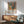 May include: Framed colorful art print featuring stylized vases with flowers, fruits, and botanical elements in vibrant oranges, greens, and blues, displayed above a wooden sideboard with white ceramic lamp. Mid-century modern living room setting with mint green upholstered armchair, round coffee table, and natural sunlight casting shadows on white wall.
