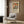 May include: Colorful floral art print in light wooden frame hanging on wood-paneled wall above beige upholstered armchair with geometric patterned pillow. Artwork features vibrant botanical design with orange, pink, green and blue flowers and leaves. Modern reading nook with brass floor lamp, dark round side table with open book, and light wood flooring.