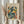 May include: Person in cream sweater holding framed art poster featuring abstract cut-out style figures in blue, teal, yellow, orange and red shapes on beige background. Light wood frame, dried pampas grass visible to left, soft natural lighting against white curtained window.