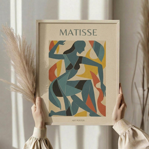 May include: Person in cream sweater holding framed art poster featuring abstract cut-out style figures in blue, teal, yellow, orange and red shapes on beige background. Light wood frame, dried pampas grass visible to left, soft natural lighting against white curtained window.