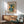 May include: Framed art poster featuring abstract cut-out style figures in teal, orange, and yellow geometric shapes on cream background, displayed above wooden sideboard with white ceramic lamp, next to mint green mid-century armchair with wooden arms, creating modern living room scene with natural sunlight casting shadows on white wall.