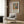 May include: Modern living room setting with beige upholstered armchair featuring geometric patterned pillow, gold floor lamp, dark round side table with open book, and wood-paneled wall. Framed poster displays colorful still life artwork with vases, plants, and fruit in vibrant oranges, greens, and blues alongside exhibition text about Matisse's artistic style and color mastery.